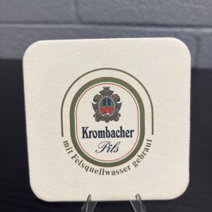 Vintage Krombacher Pils beer coaster, featuring the brewery's heritage story and crest, a high-vibration collectible perfect for enthusiasts of international breweriana looking to manifest prosperity and add authentic brand history to their portfolio.