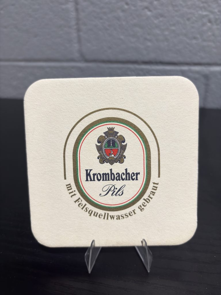 Vintage Krombacher Pils beer coaster, featuring the brewery's heritage story and crest, a high-vibration collectible perfect for enthusiasts of international breweriana looking to manifest prosperity and add authentic brand history to their portfolio.