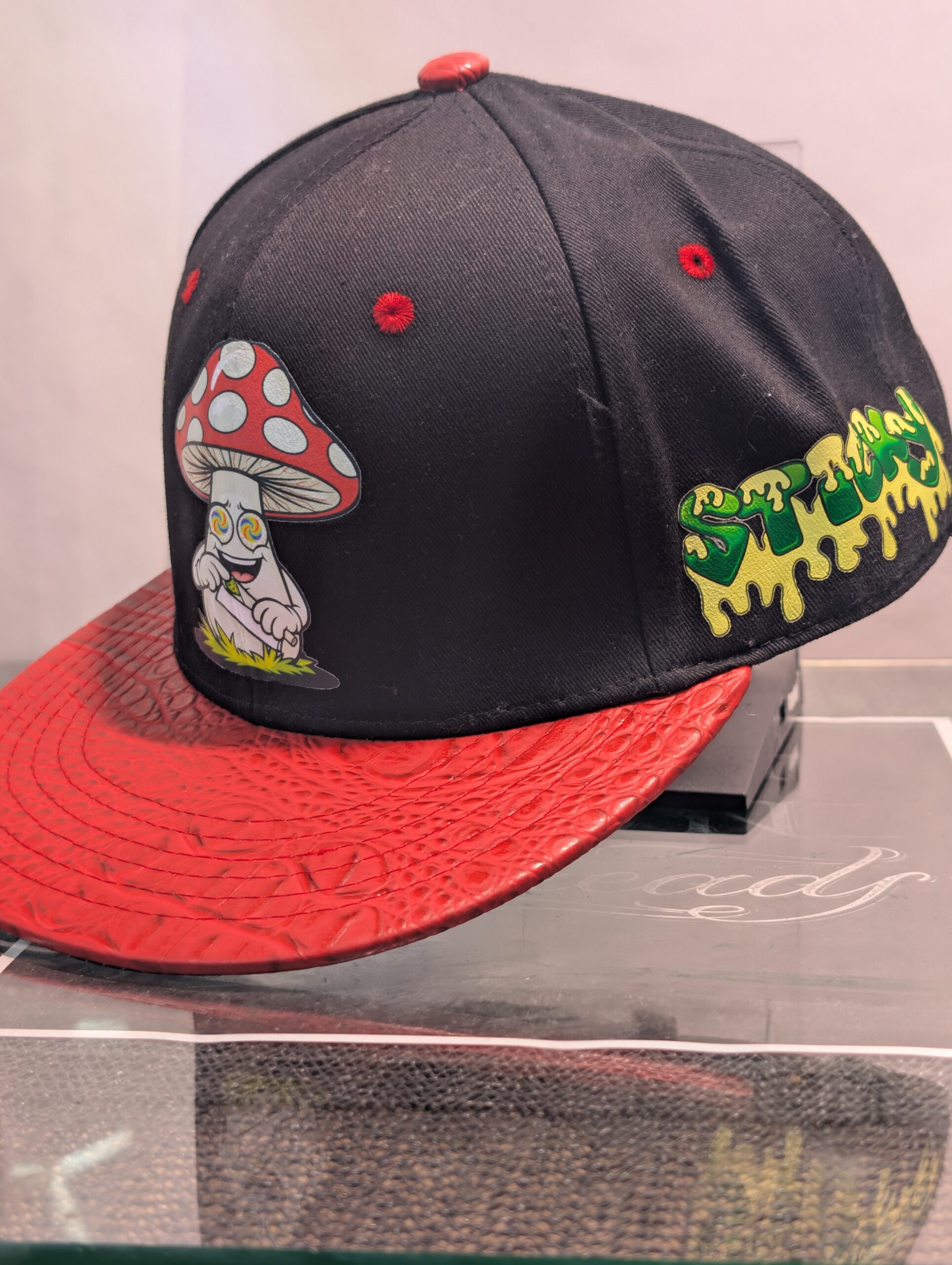 Black structured streetwear snapback hat featuring embroidered cartoon mushroom graphic alongside bright green dripping slime logo and textured red crocodile faux leather flat bill edge for style.