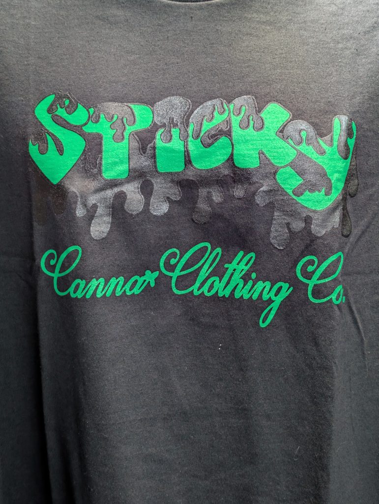 Black shirt featuring green Sticky Canna drip logo.