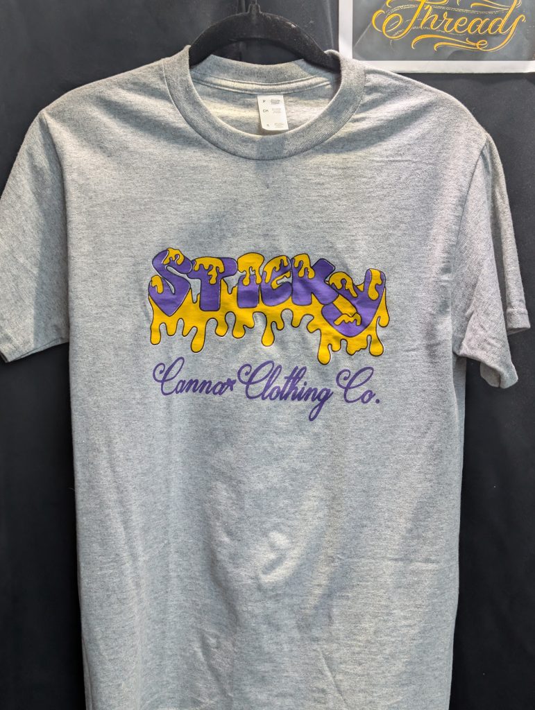 Grey shirt featuring purple yellow Sticky Canna logo.
