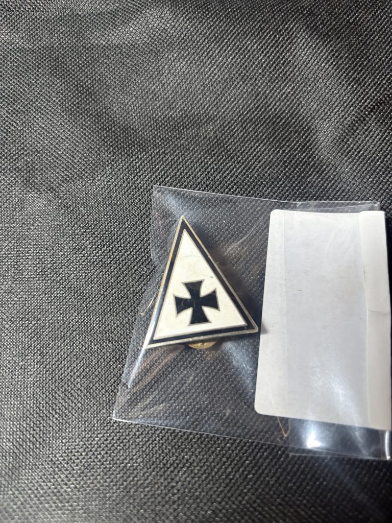 Vintage triangular lapel pin with black cross emblem on white background, in a protective sleeve.