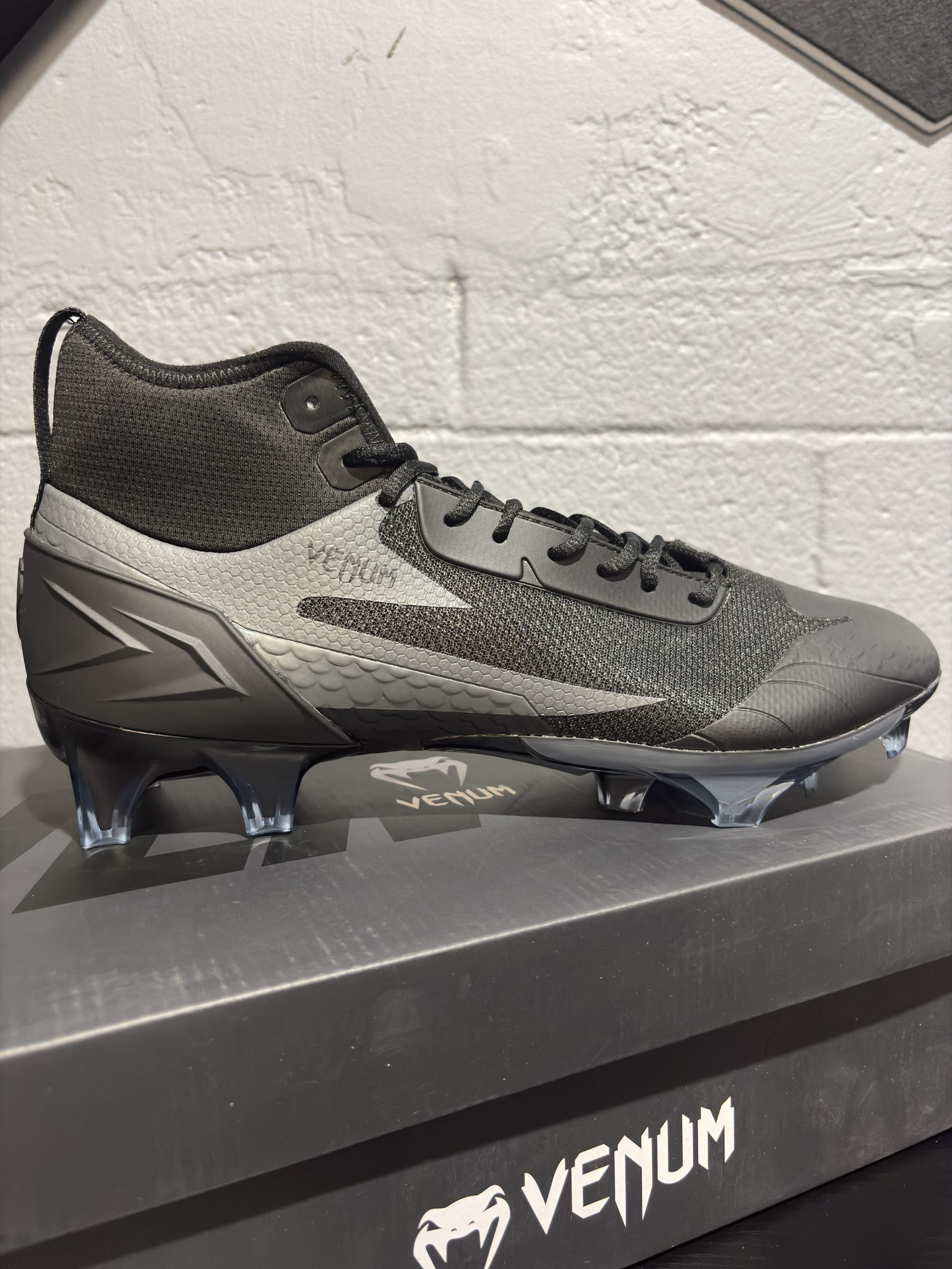 Venum Elite Football Cleats in Black/Black displayed next to their branded shoebox.