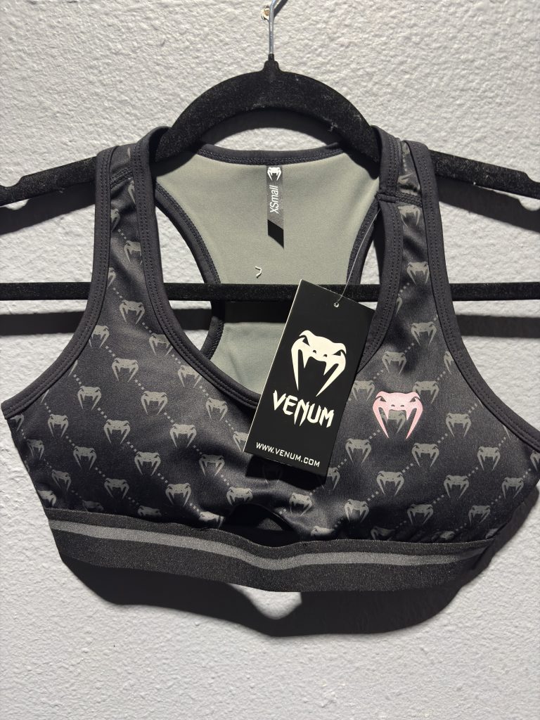 Venum Monogram Sports Bra in Black and Pink Gold showing the repeating logo pattern and hanging retail tag in size XS.