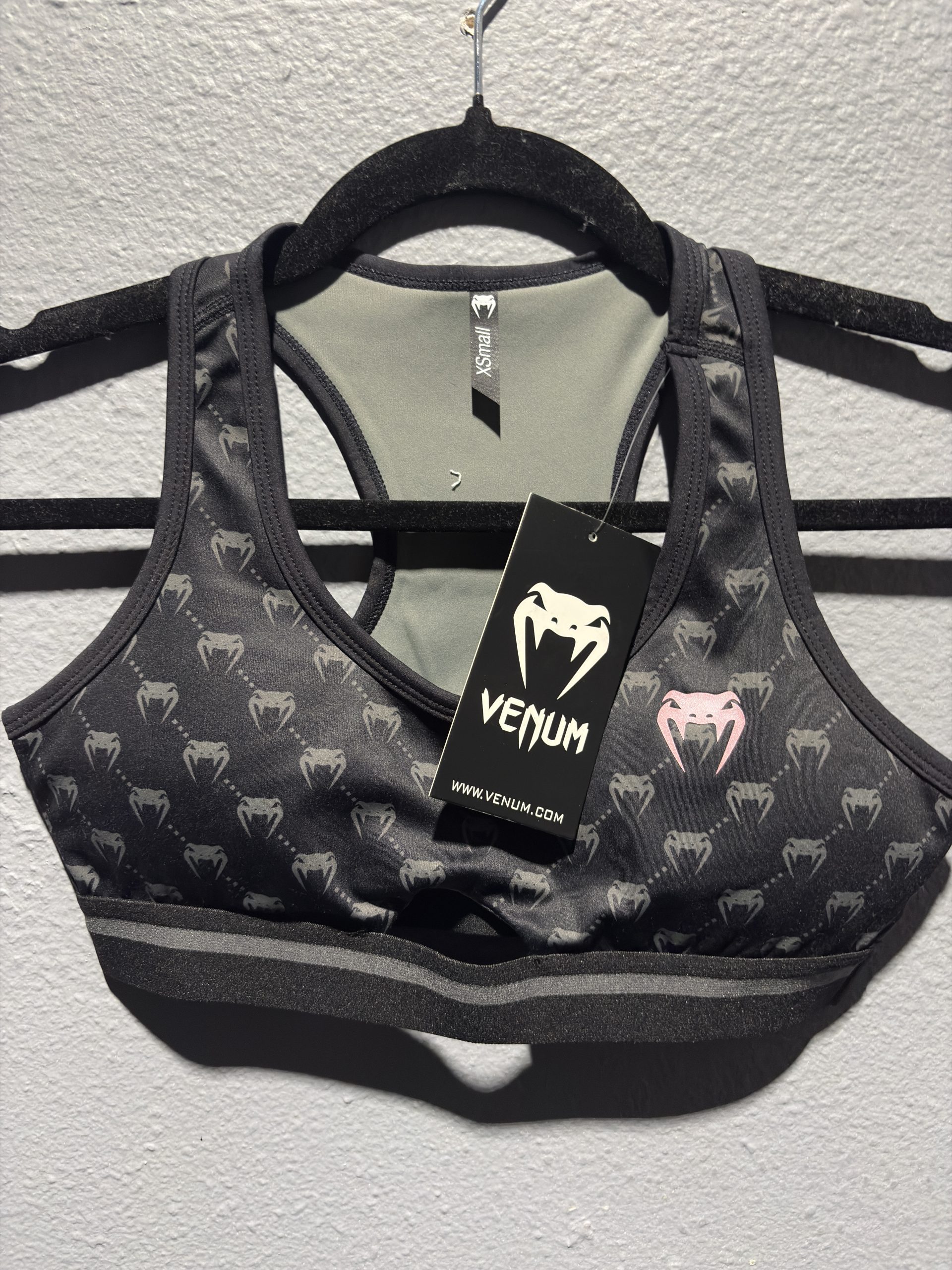 Venum Monogram Sports Bra in Black and Pink Gold showing the repeating logo pattern and hanging retail tag in size XS.