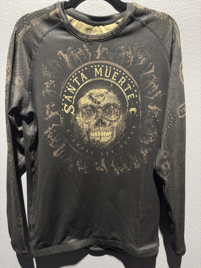 Venum Santa Muerte Rashguard in Black and Brown showing the front chest graphic, collar detailing, and retail tag in size 2XL.