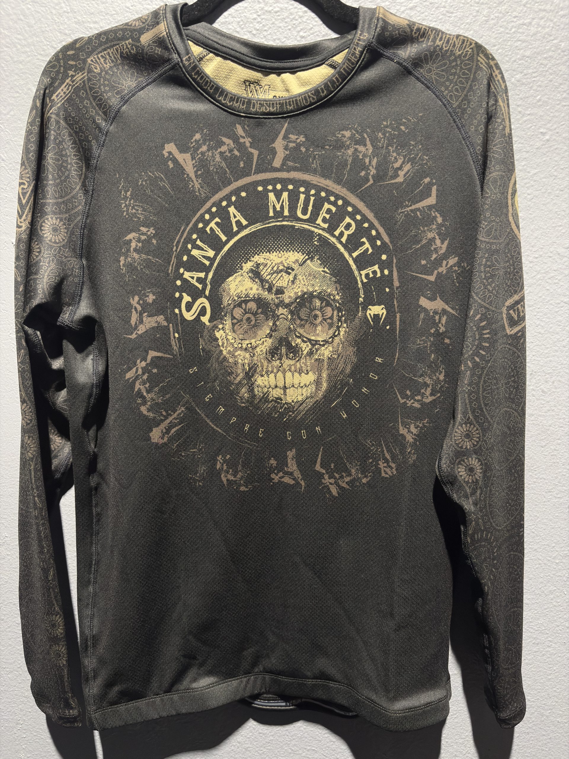 Venum Santa Muerte Rashguard in Black and Brown showing the front chest graphic, collar detailing, and retail tag in size 2XL.