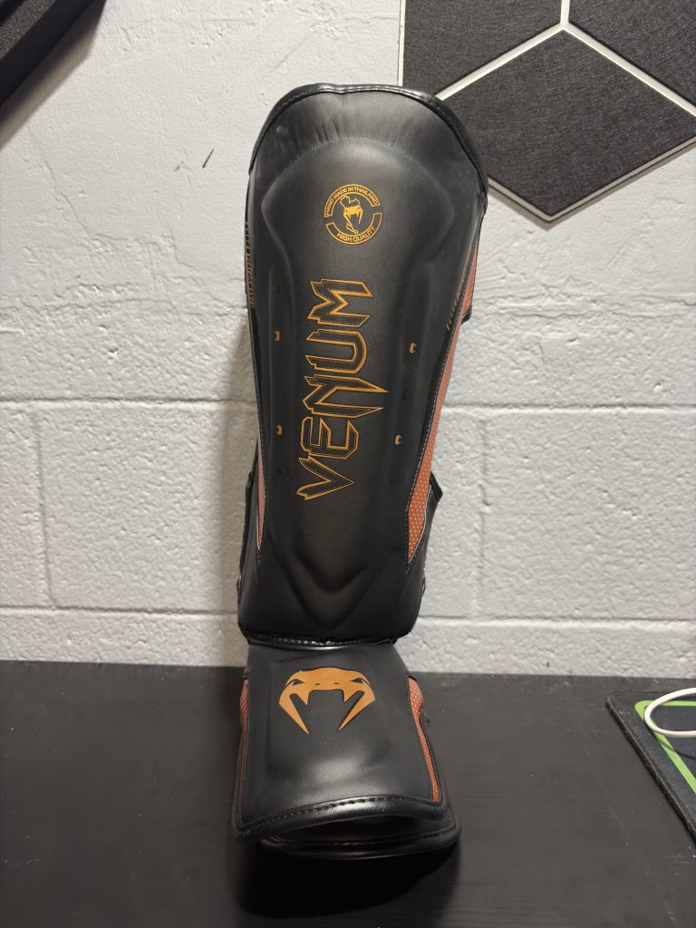 Venum Elite Shin Guards in Black and Bronze showing the front shin logo, side perforated panels, and back closure straps.