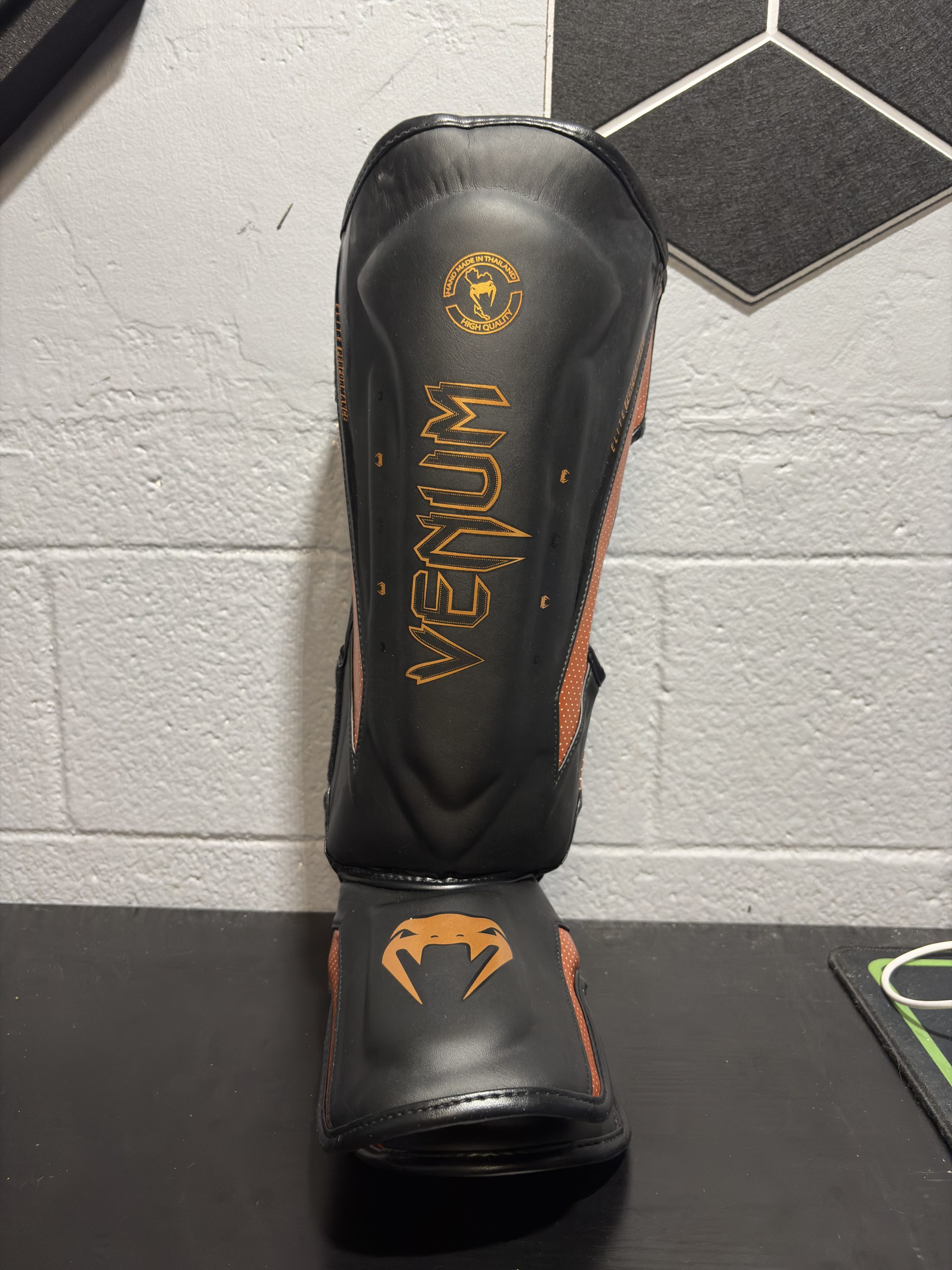 Venum Elite Shin Guards in Black and Bronze showing the front shin logo, side perforated panels, and back closure straps.