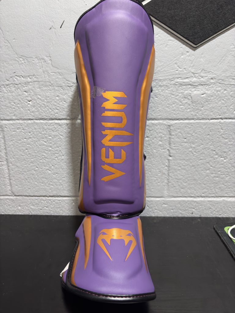 Venum Elite Shin Guards in Purple and Gold showing the front shin logo, side metallic gold panels, and size L back strap.