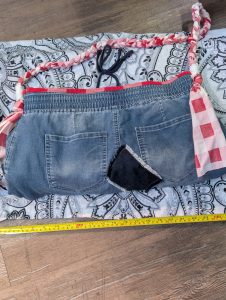 Upcycled distressed denim shoulder bag with red plaid trim, patchwork accents, and pink braided shoulder strap shown with a measuring tape.