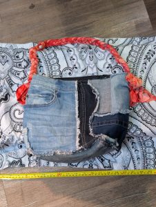 Upcycled patchwork denim shoulder bag with raw edges, mixed wash denim panels, and red ombre braided strap shown with a measuring tape.