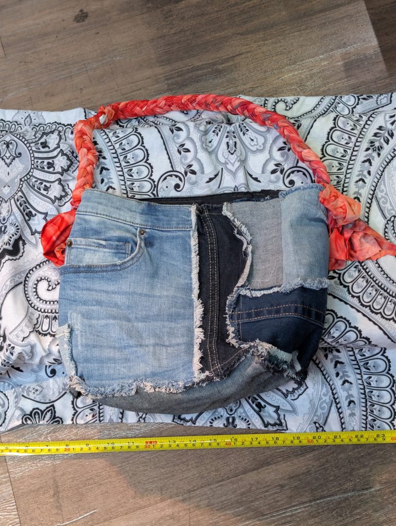 Upcycled patchwork denim shoulder bag with raw edges, mixed wash denim panels, and red ombre braided strap shown with a measuring tape.