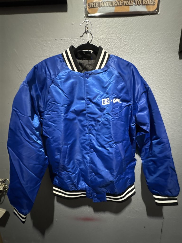 Shaka Wear The Art and Wellness Club satin varsity jacket in royal blue showing the front chest logo, back script graphic, and hanging tag in size S.